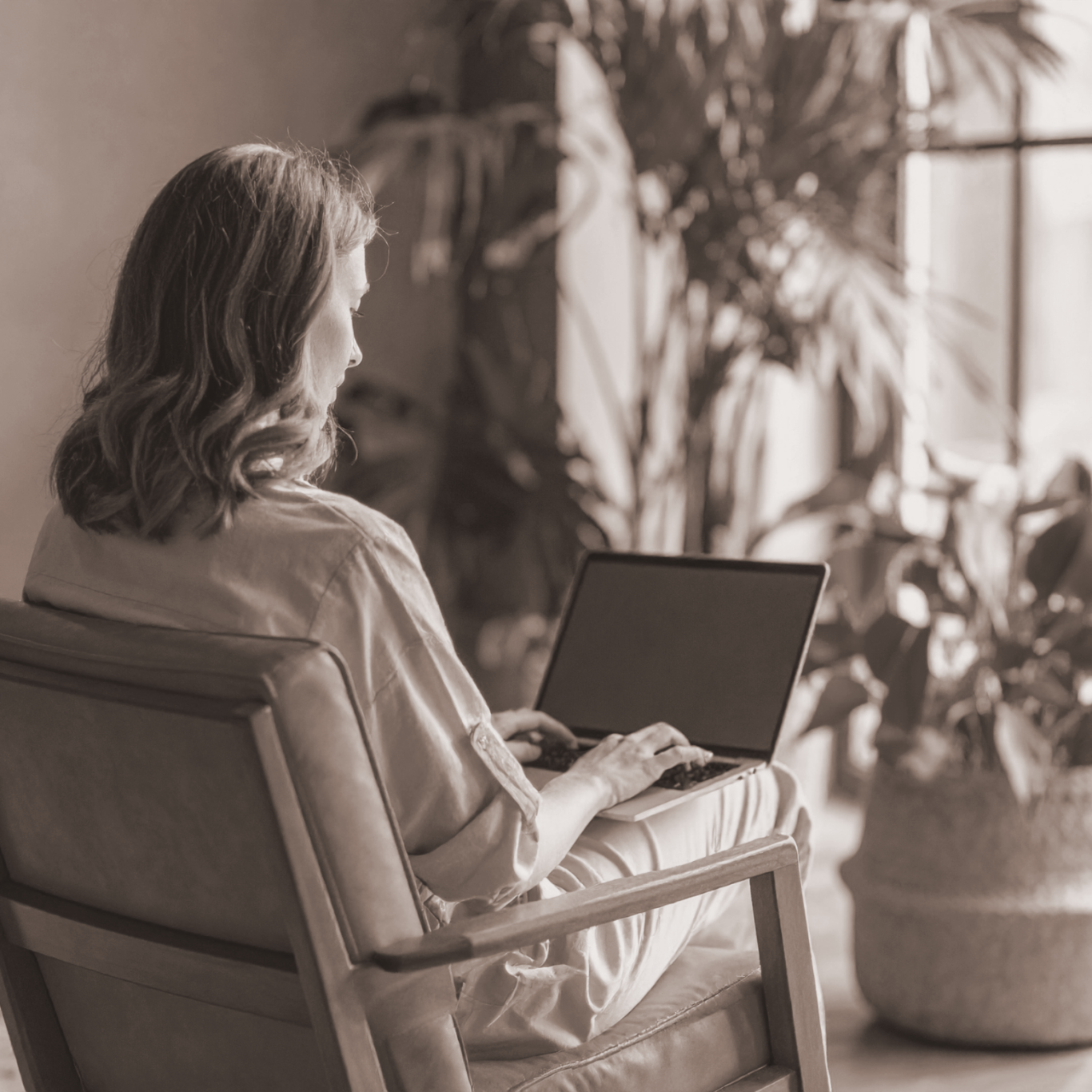 Frau mit Locken sitzt auf einem Sessel und arbeitet am Laptop, umgeben von Pflanzen.